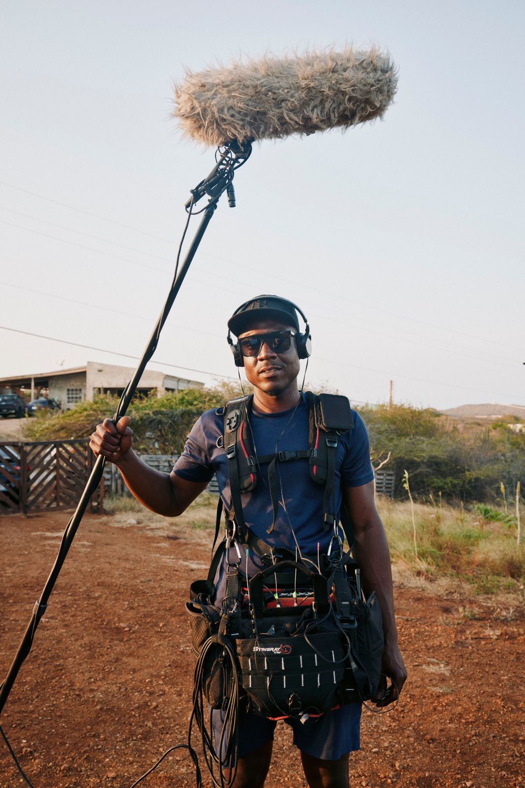 Sound technician with boom pole and gear standing outdoors in rural area.