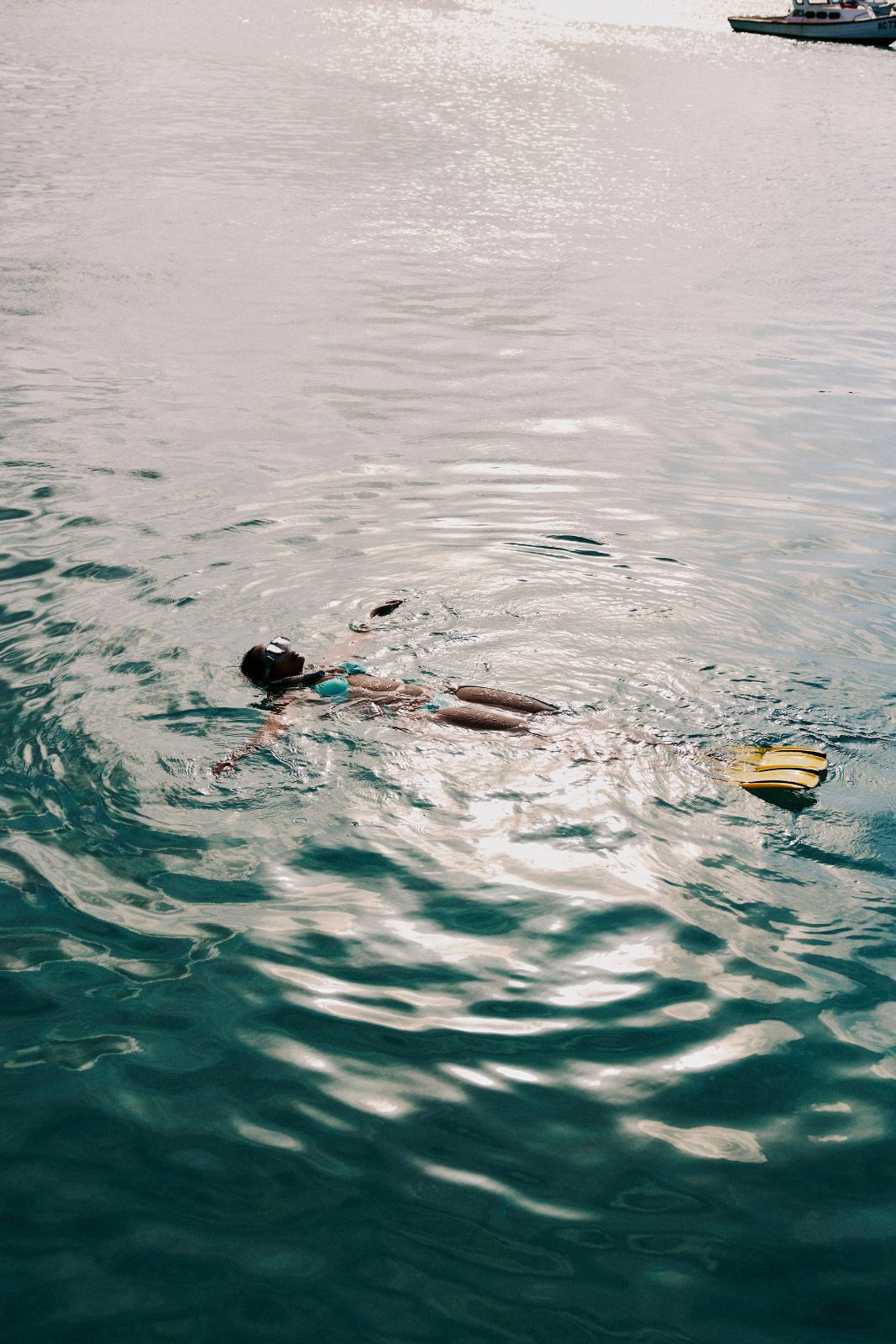 Snorkeler floating on their back in shimmering ocean water with distant boat.