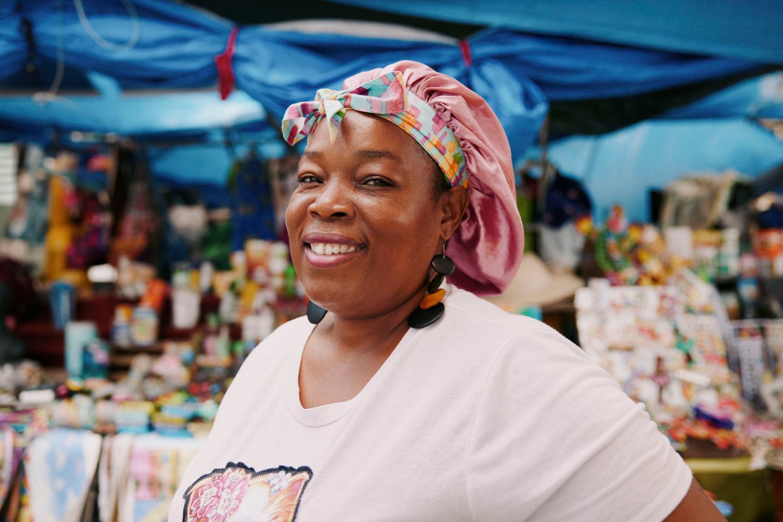 Smiling woman in pink headwrap at vibrant outdoor Curaçao market stall.