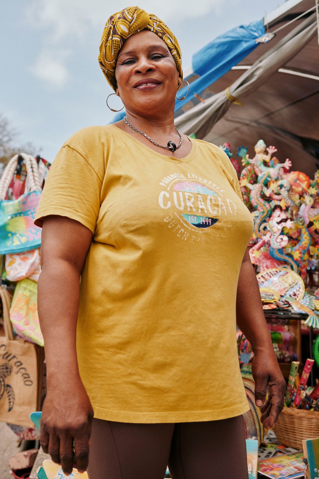 Smiling woman in yellow shirt stands by colorful Curaçao souvenirs outdoors.