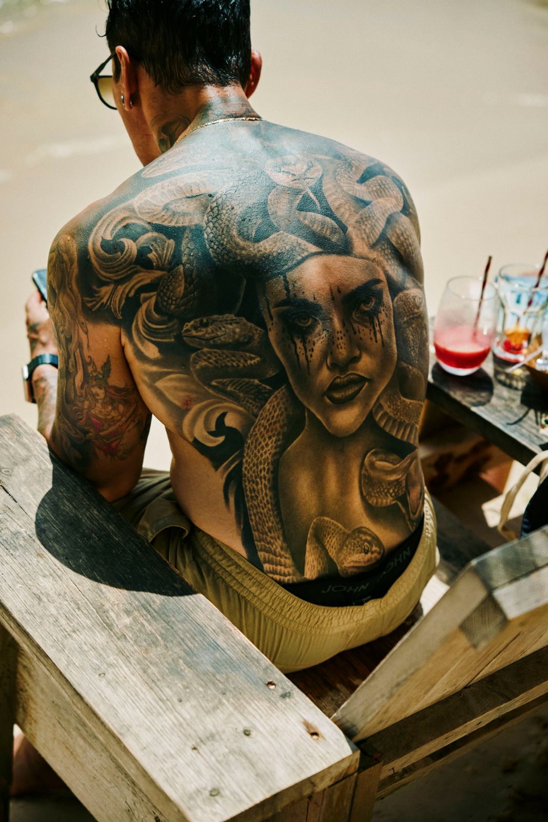 Back tattoo of Medusa on a man sitting near colorful drinks outdoors.