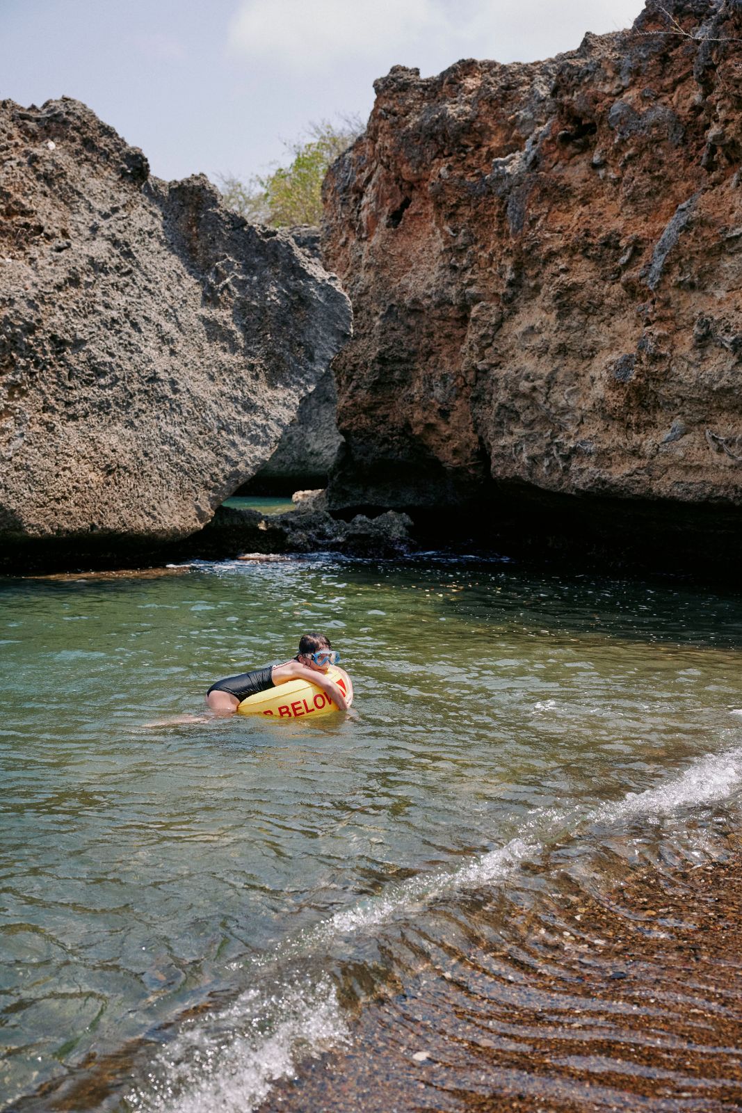 Child with goggles floating on a yellow buoy near rocky coastal formations.