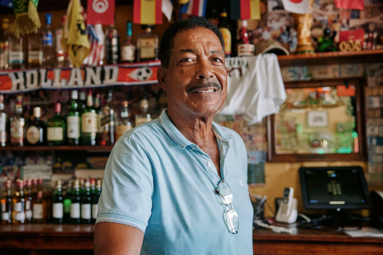 Smiling local man in light blue polo behind cluttered, flag-draped bar.