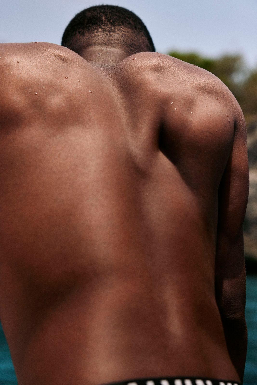 Muscular sun-drenched back of a man sitting outdoors near the water.