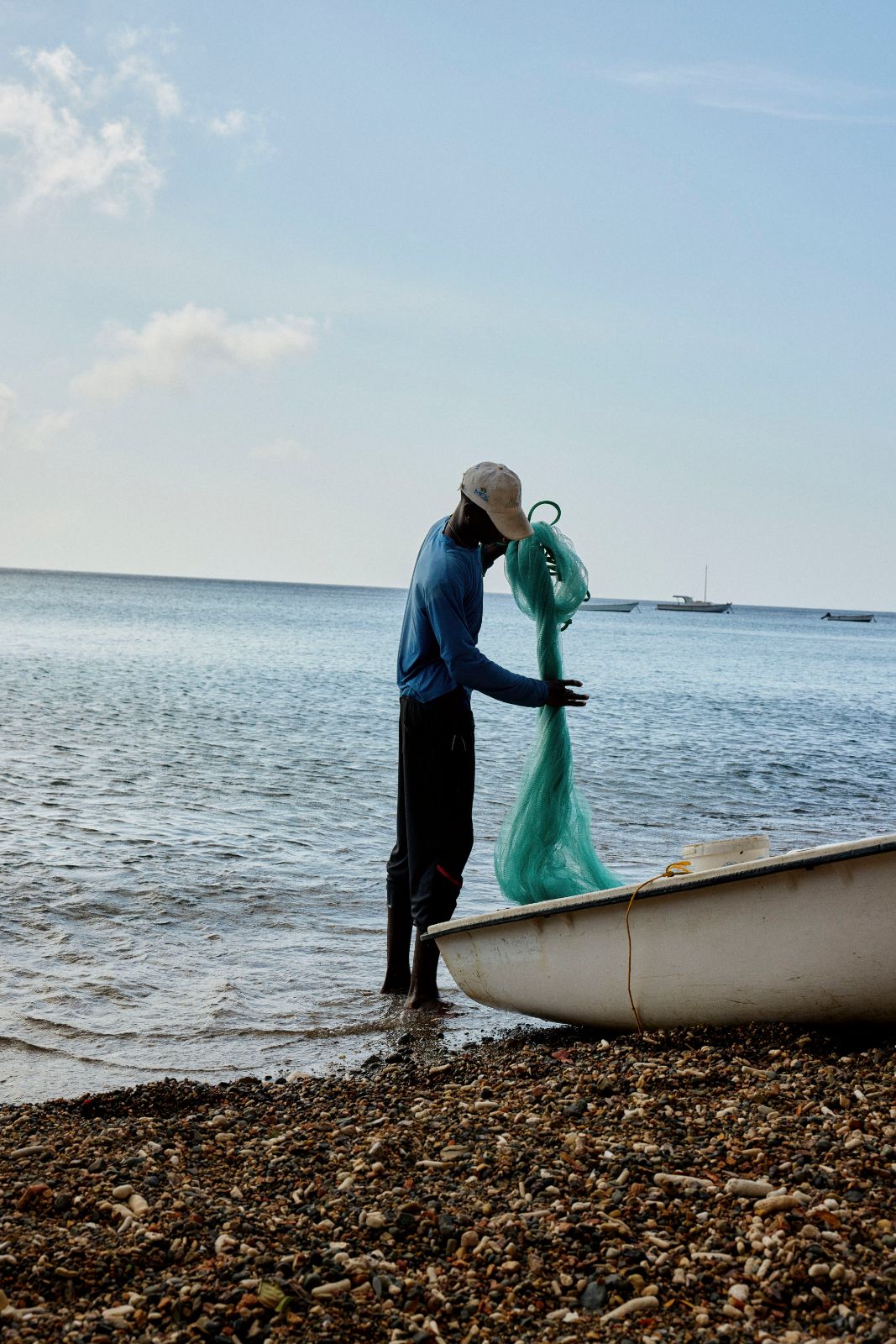 Fisherman gathering green fishing net beside a white boat on the shore.