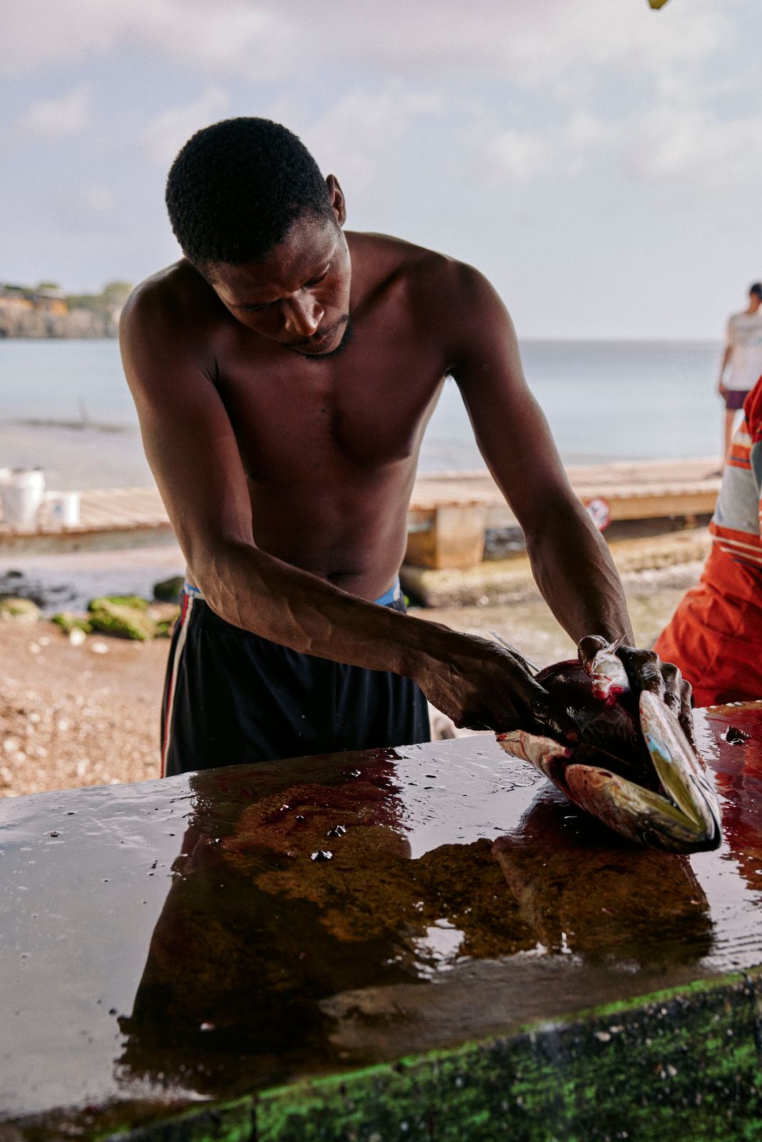 Shirtless fisherman meticulously gutting a large fish on a wet table.