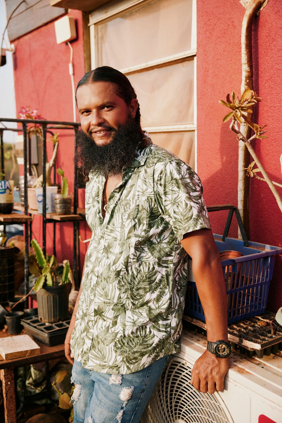 Smiling bearded man in tropical shirt leaning against a red building.