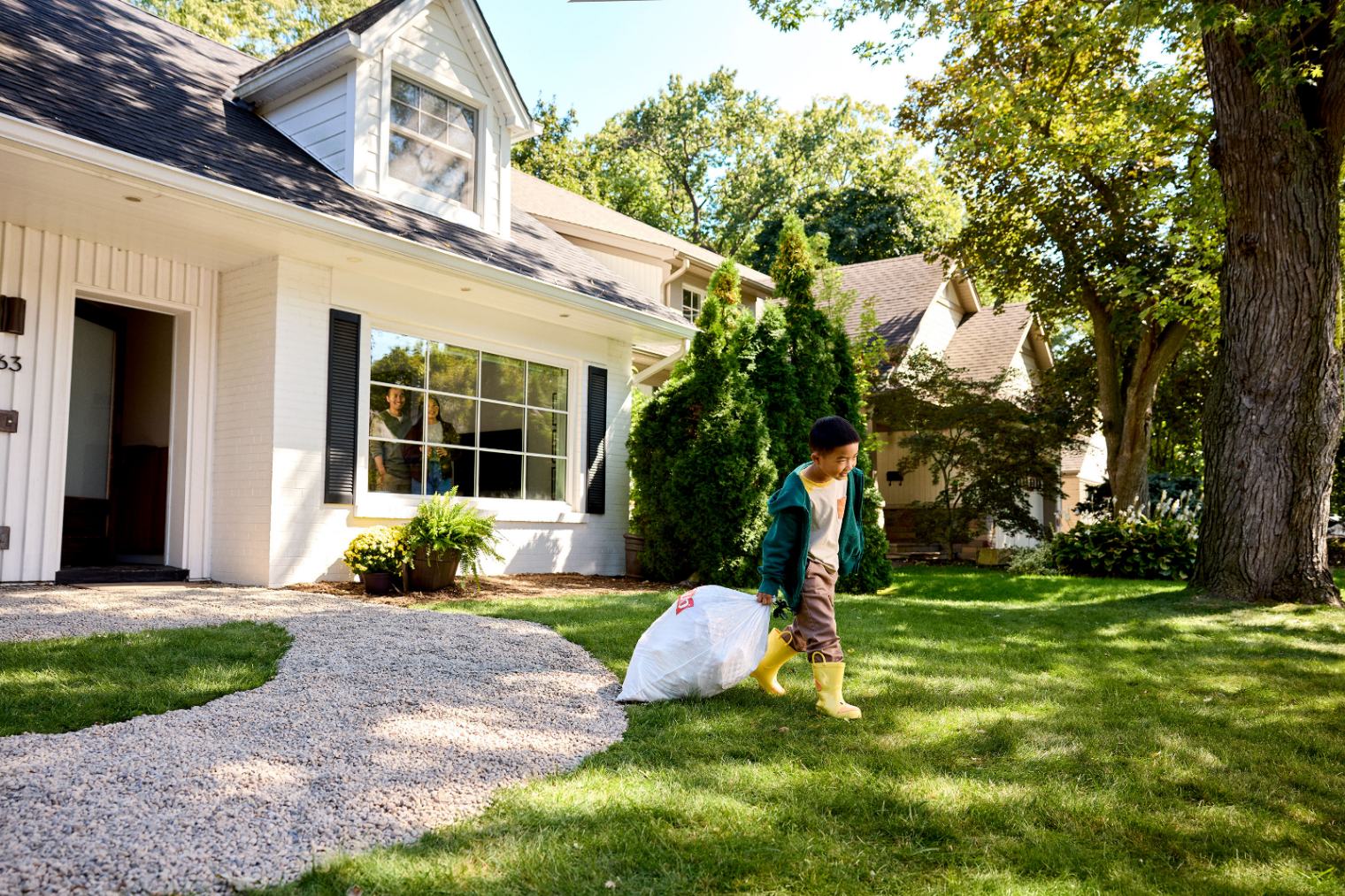 Child in yellow rain boots hauls a white trash bag across a wide suburban lawn.