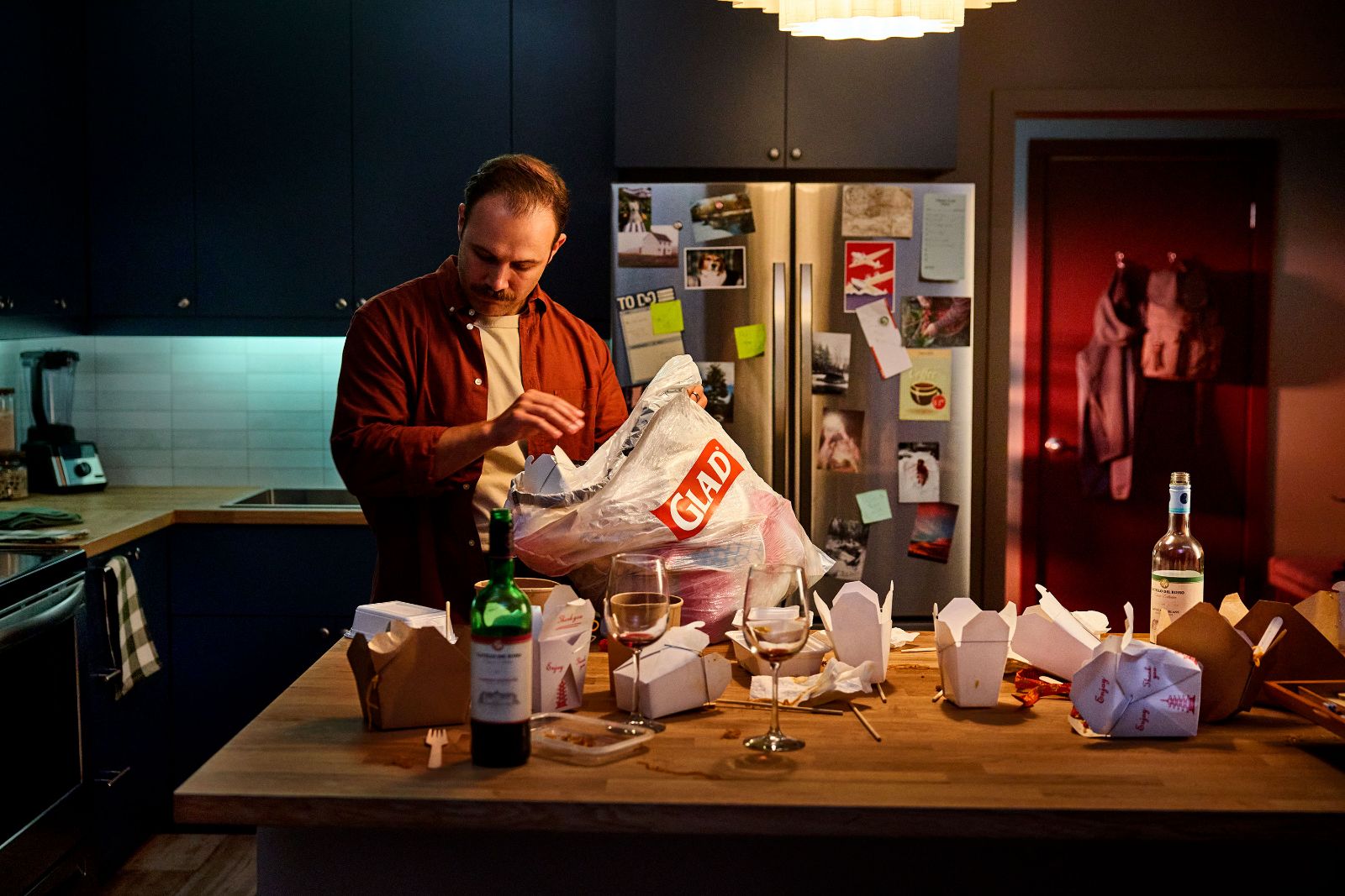 Man cleaning up takeout boxes in a dimly lit moody kitchen after a dinner party.