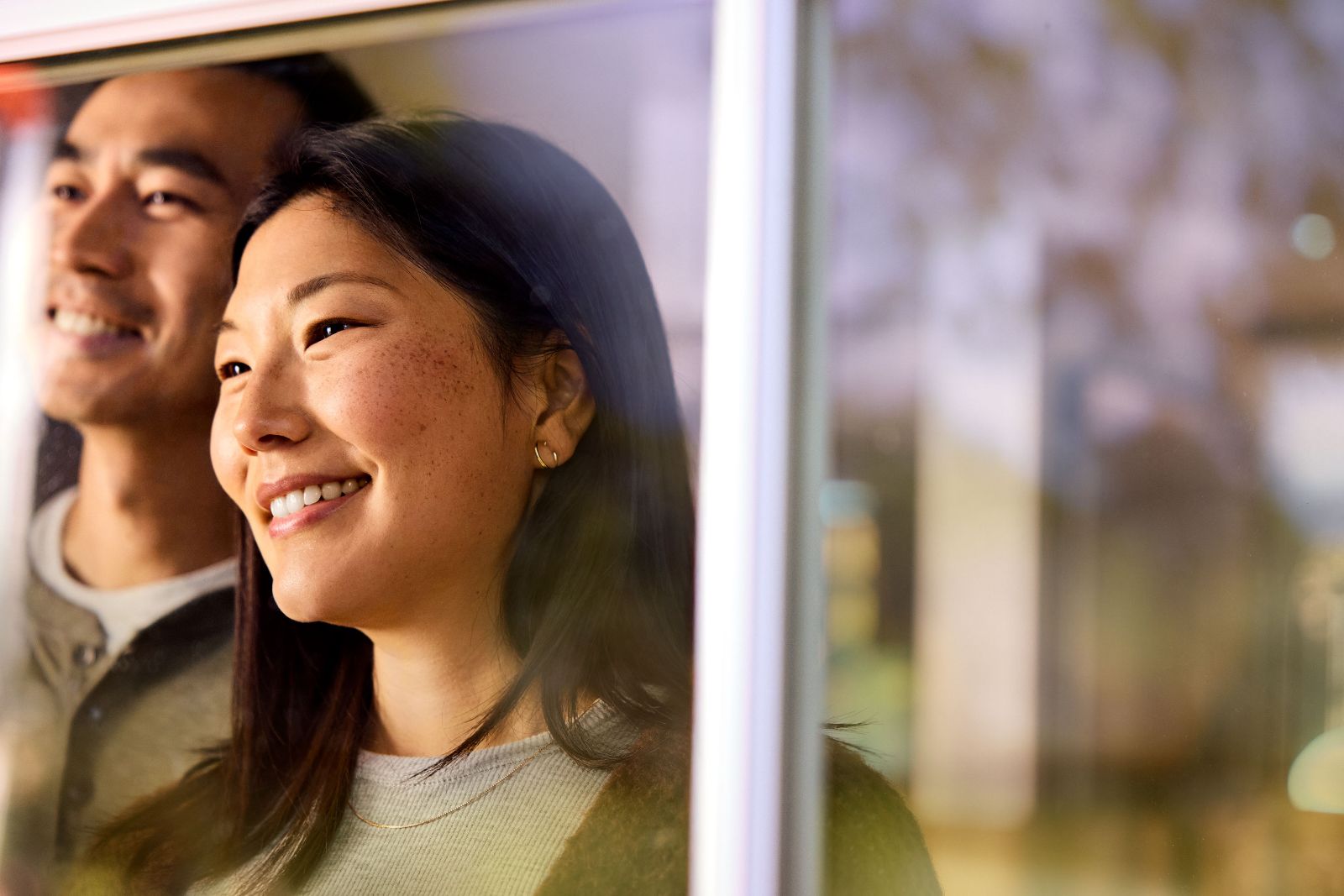Asian couple looking out a bright sunlit window reflecting the suburban oak trees outside.