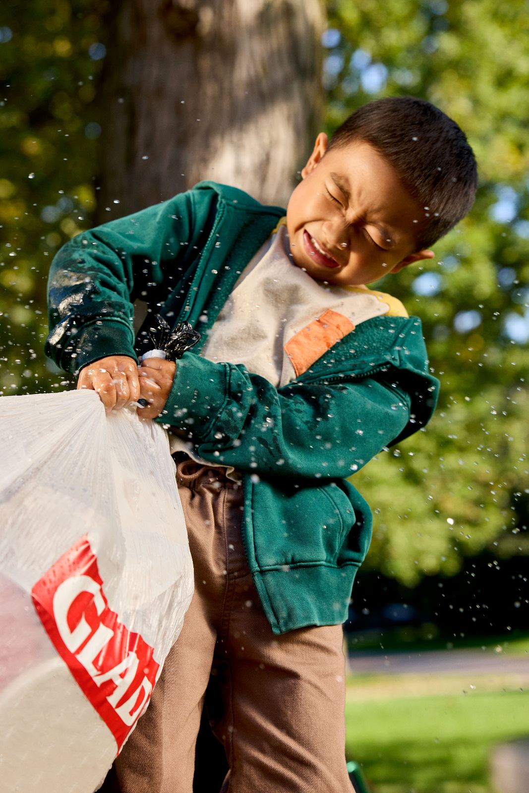 Young boy dragging a Glad garbage bag through spraying water in a sunny suburban yard.