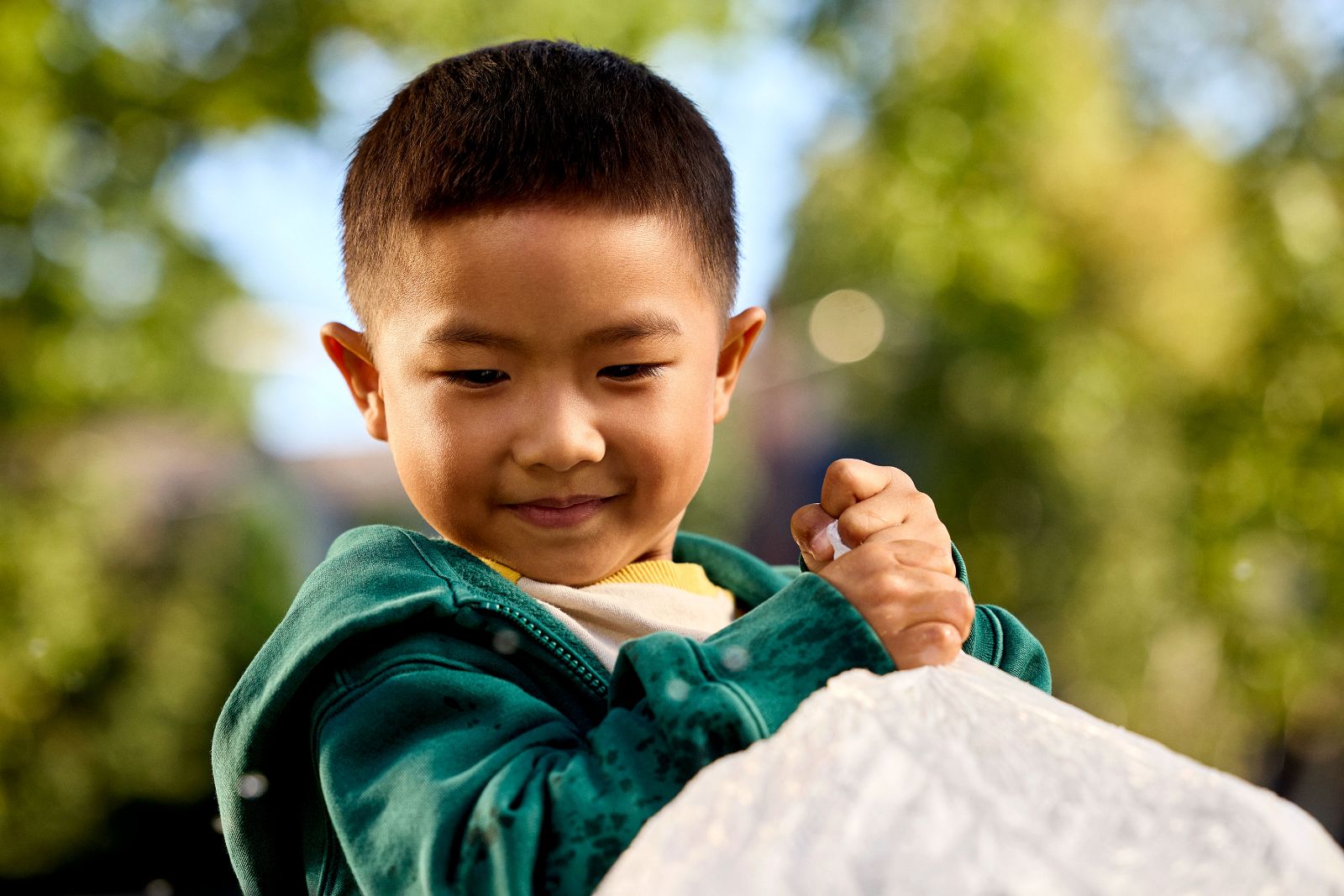 Close up of a smiling boy in a green hoodie gripping a white plastic garbage bag.