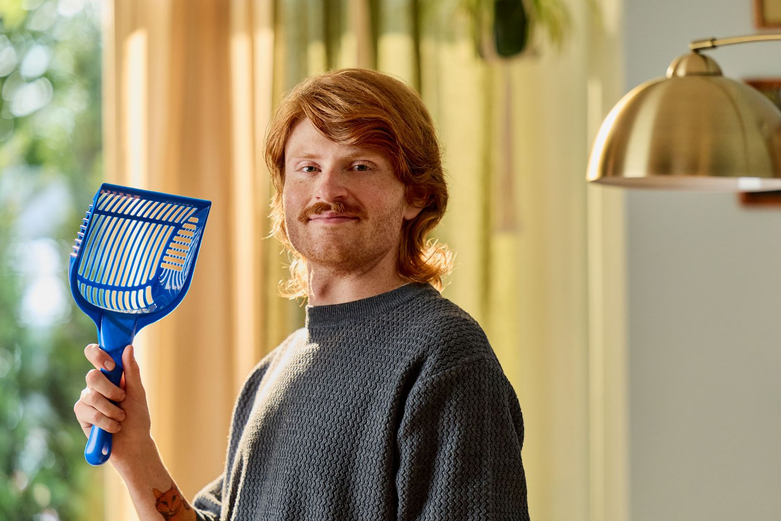 Ginger-haired man in textured sweater holding a bright blue litter scoop near sunny window.