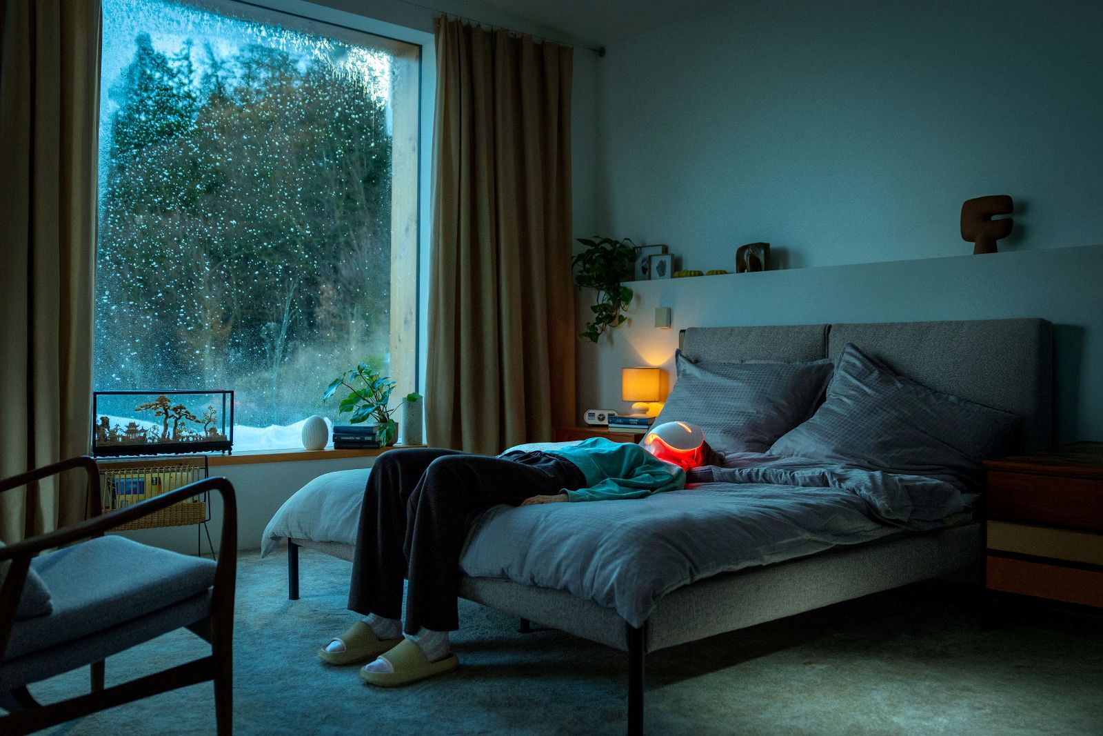 Woman lying flat on a bed in a minimalist room wearing a red LED therapy mask.