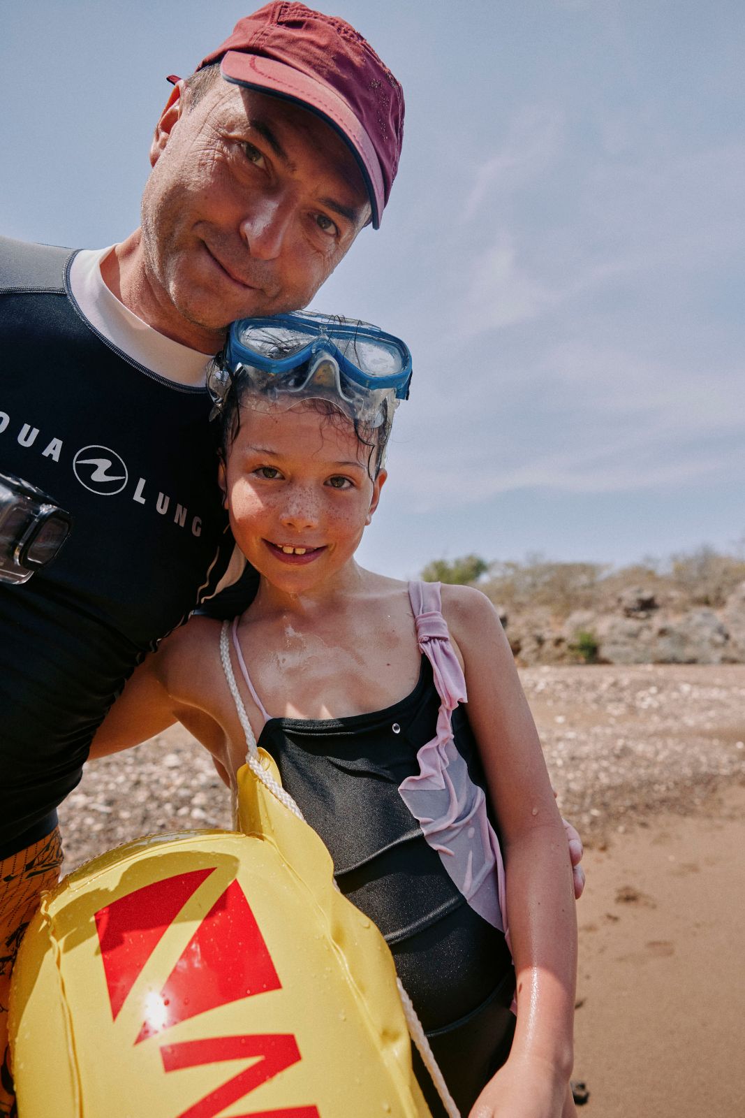 Man and young girl embracing playfully on a sandy beach under bright tropical sunshine.