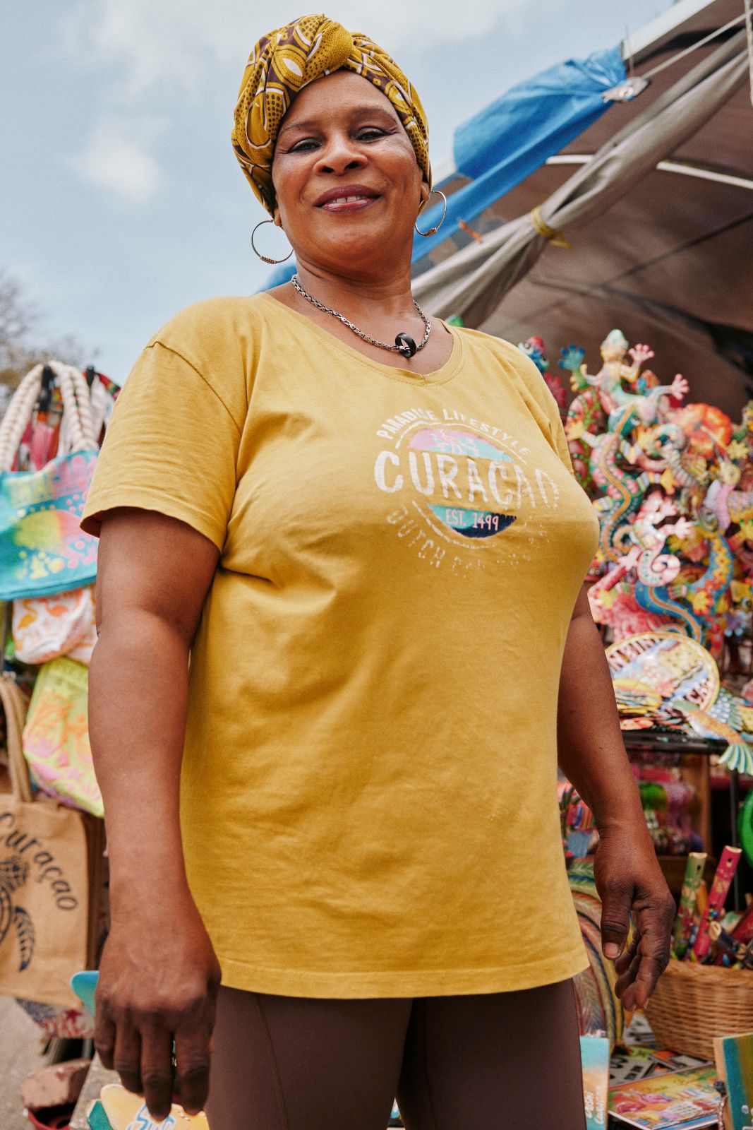 Woman wearing a mustard headwrap smiling brightly at a vibrant outdoor souvenir market.