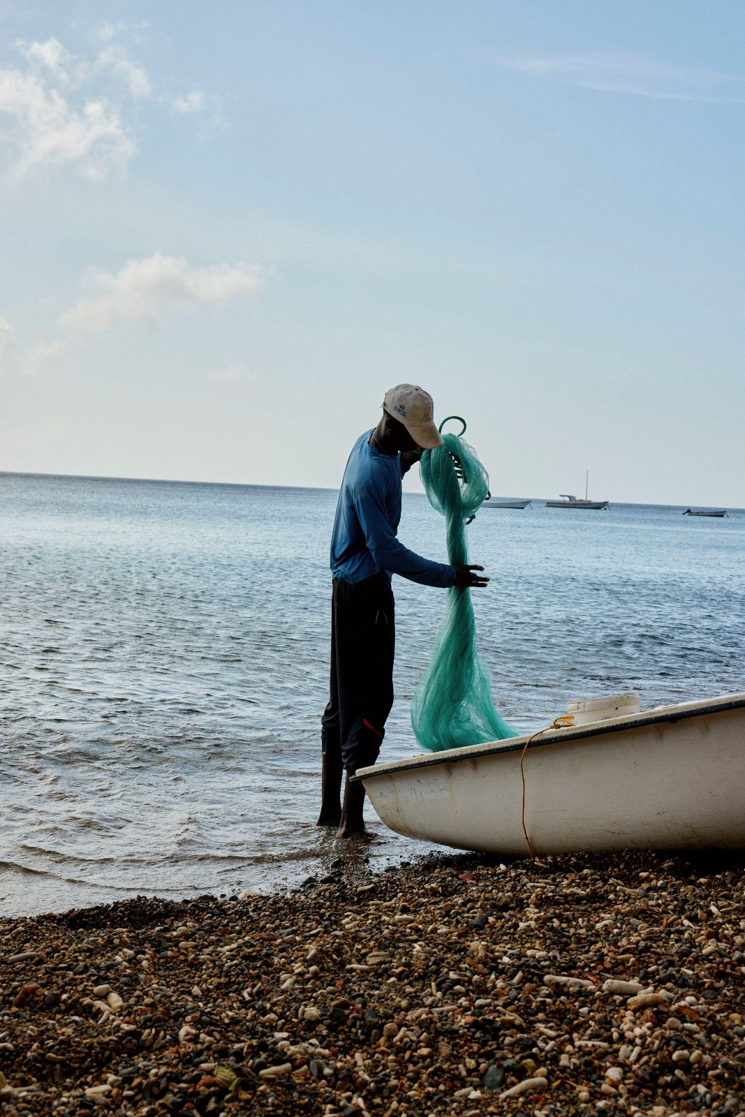 Fisherman sorting a large green net beside a white boat on a pebble beach.