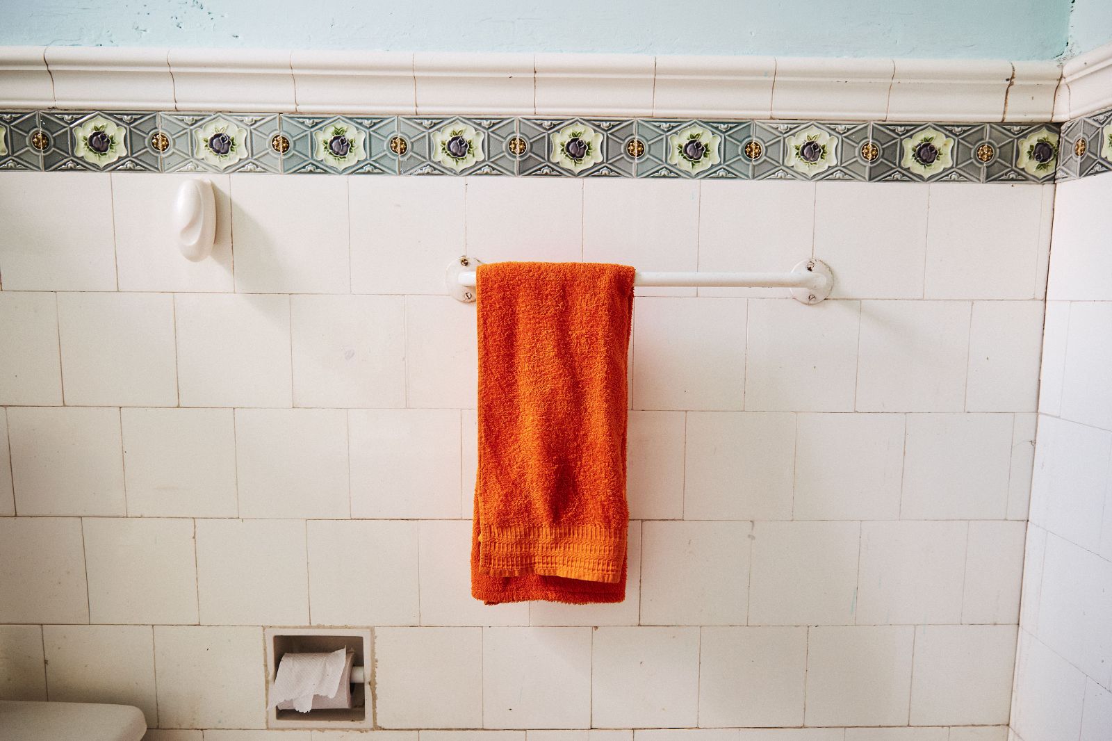 A bright orange bath towel hangs against classic white tiled walls featuring floral borders.