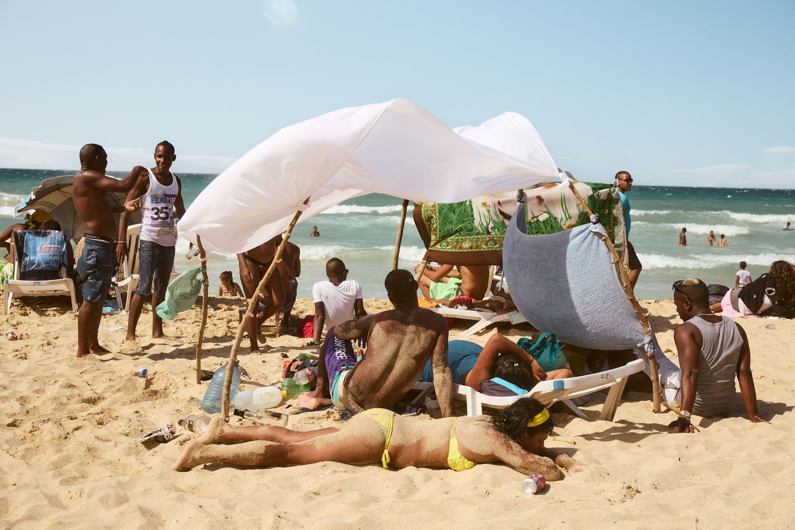 Groups of people relaxing under makeshift white cloth shade structures built on sandy shores.