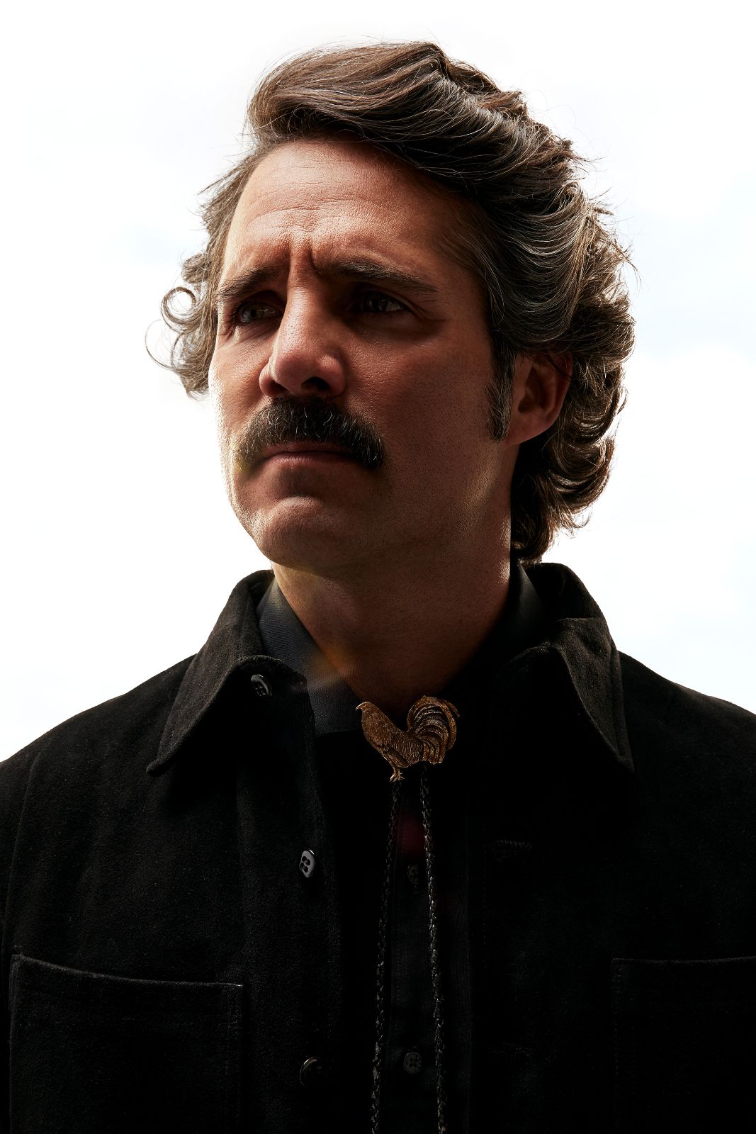 Actor Jose Ron in profile wearing a golden rooster bolo tie against a brilliant white sky.