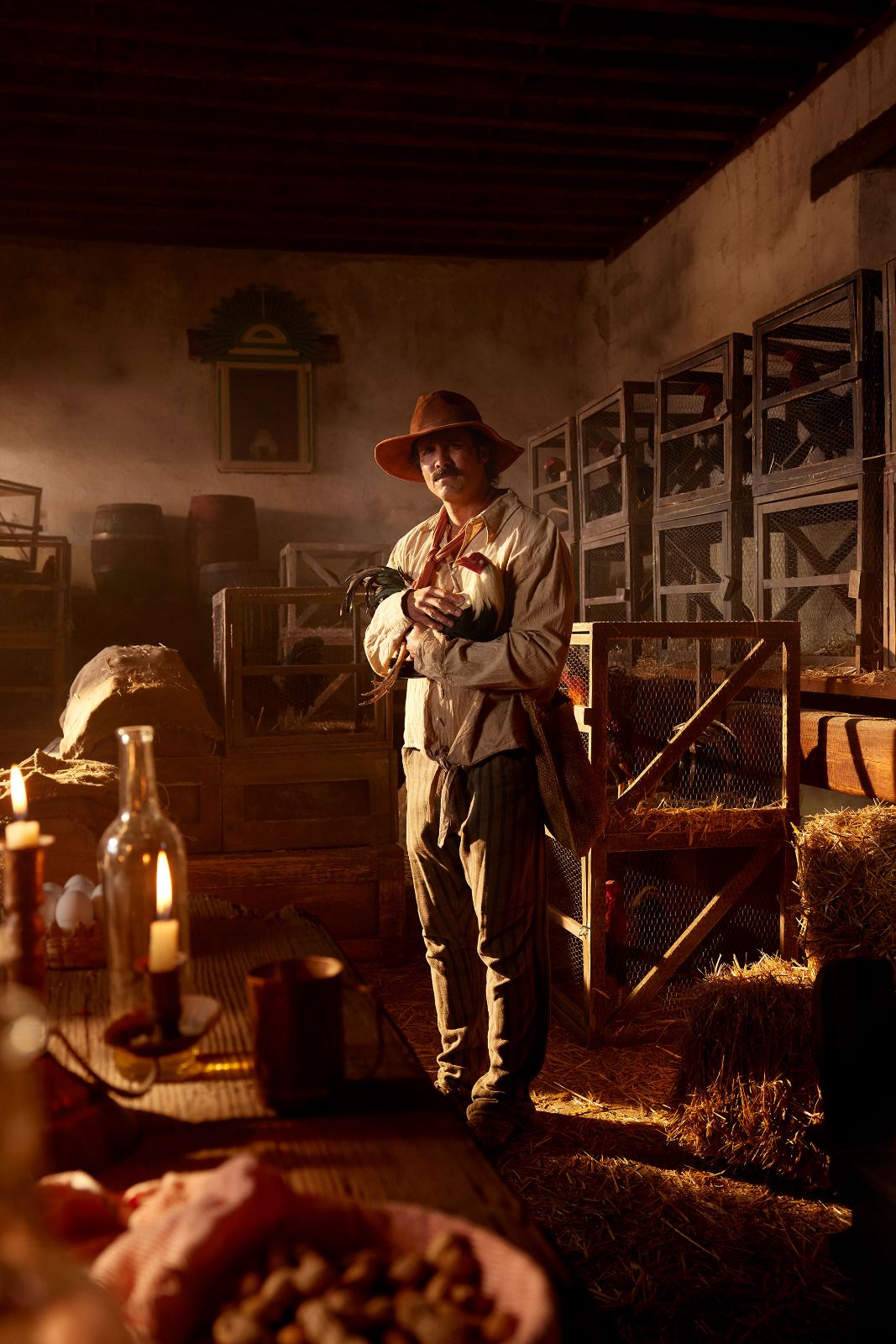 Jose Ron in a straw-filled barn, cradling a rooster near glowing candles.