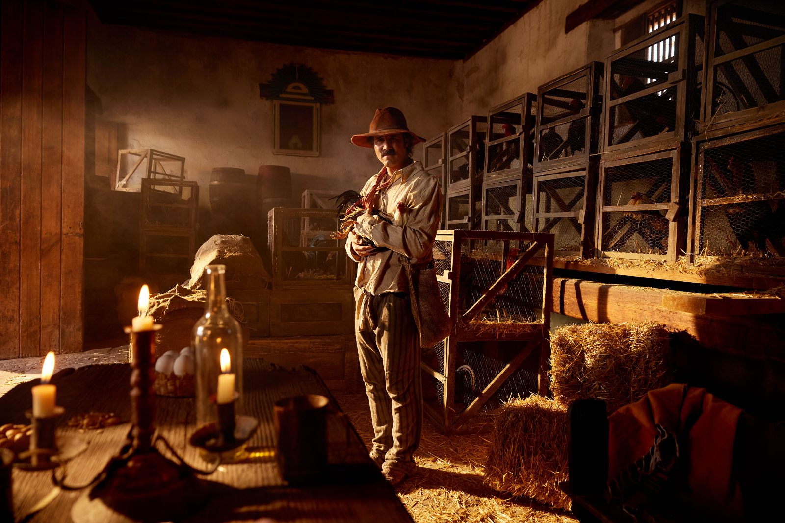 Wide view of Jose Ron in a straw-filled barn, cradling a rooster near glowing candles.