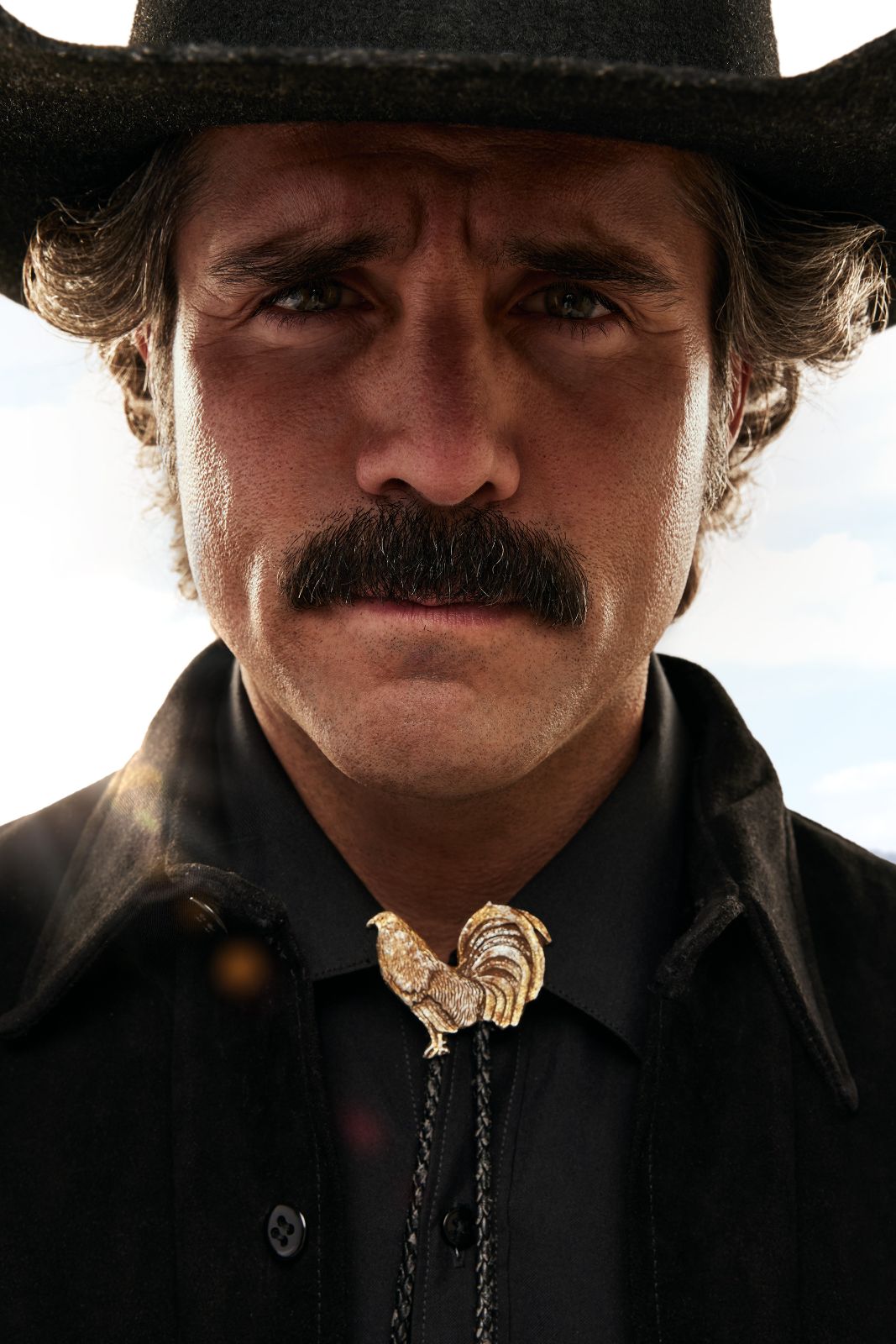 Direct, intense portrait of Jose Ron staring into the camera, wearing a rooster bolo tie.