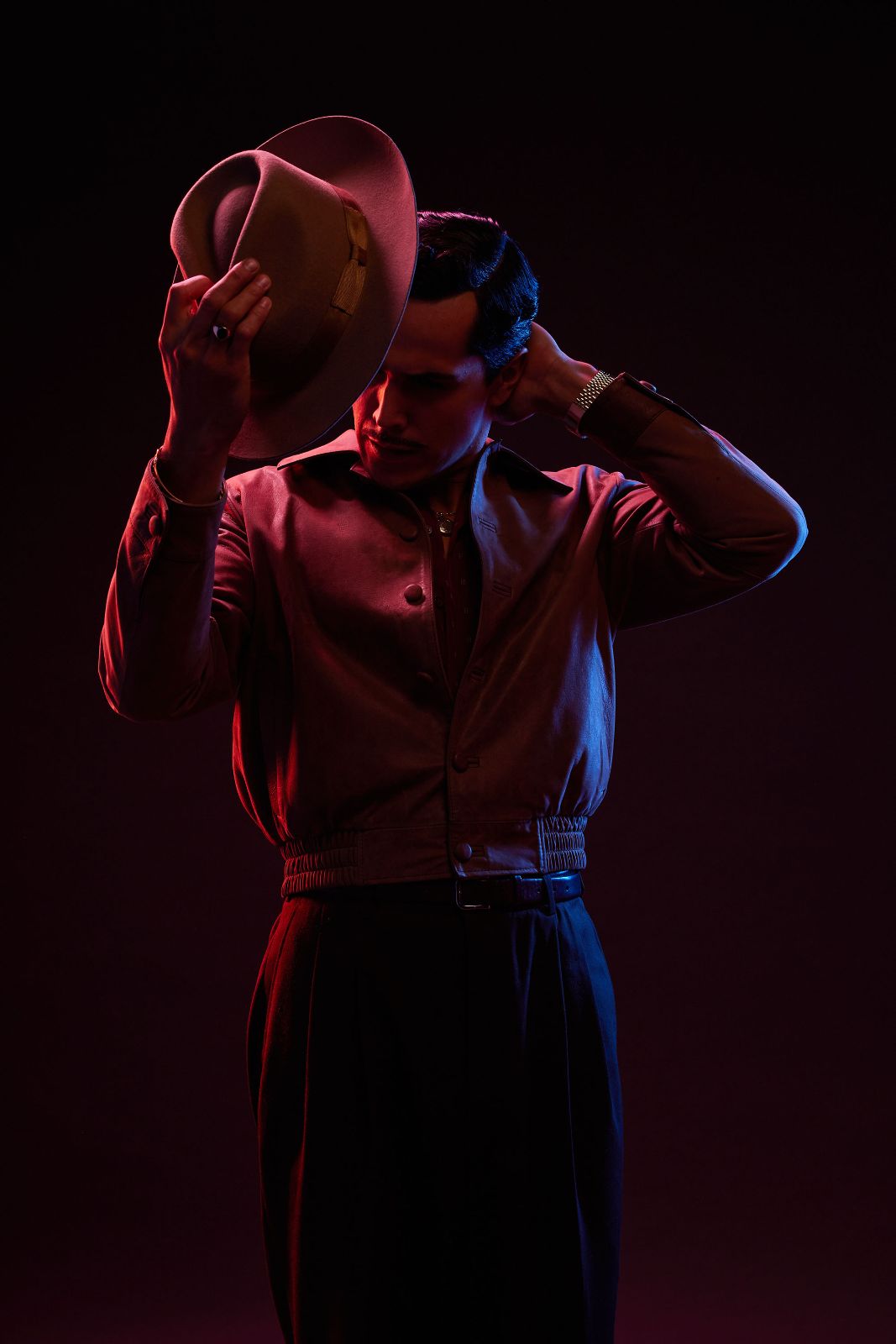 Mario Moran tipping a classic fedora downwards while bathed in dramatic red and blue studio lighting.