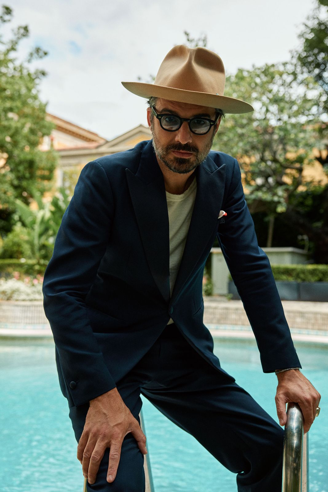 Portrait of a man in a navy suit and hat resting by a pool.