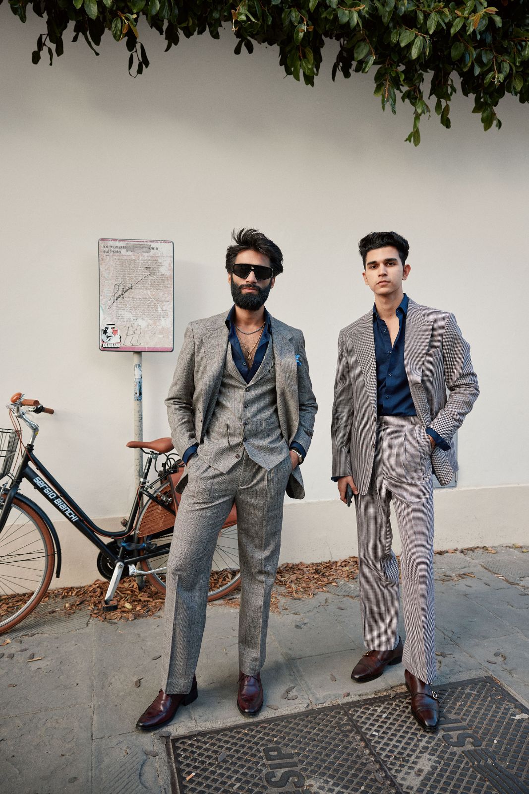 Two fashionable men posing in tailored suits against a white outdoor wall.