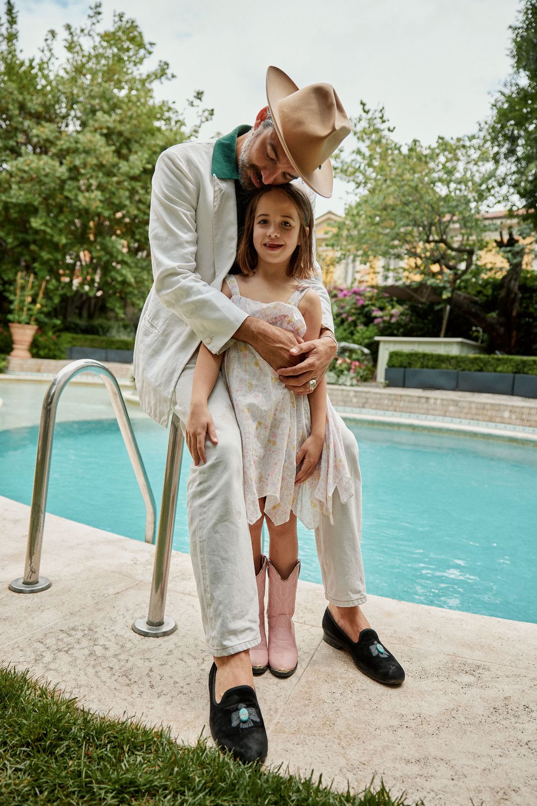 Father in a crisp white suit embraces his daughter by a lush pool.
