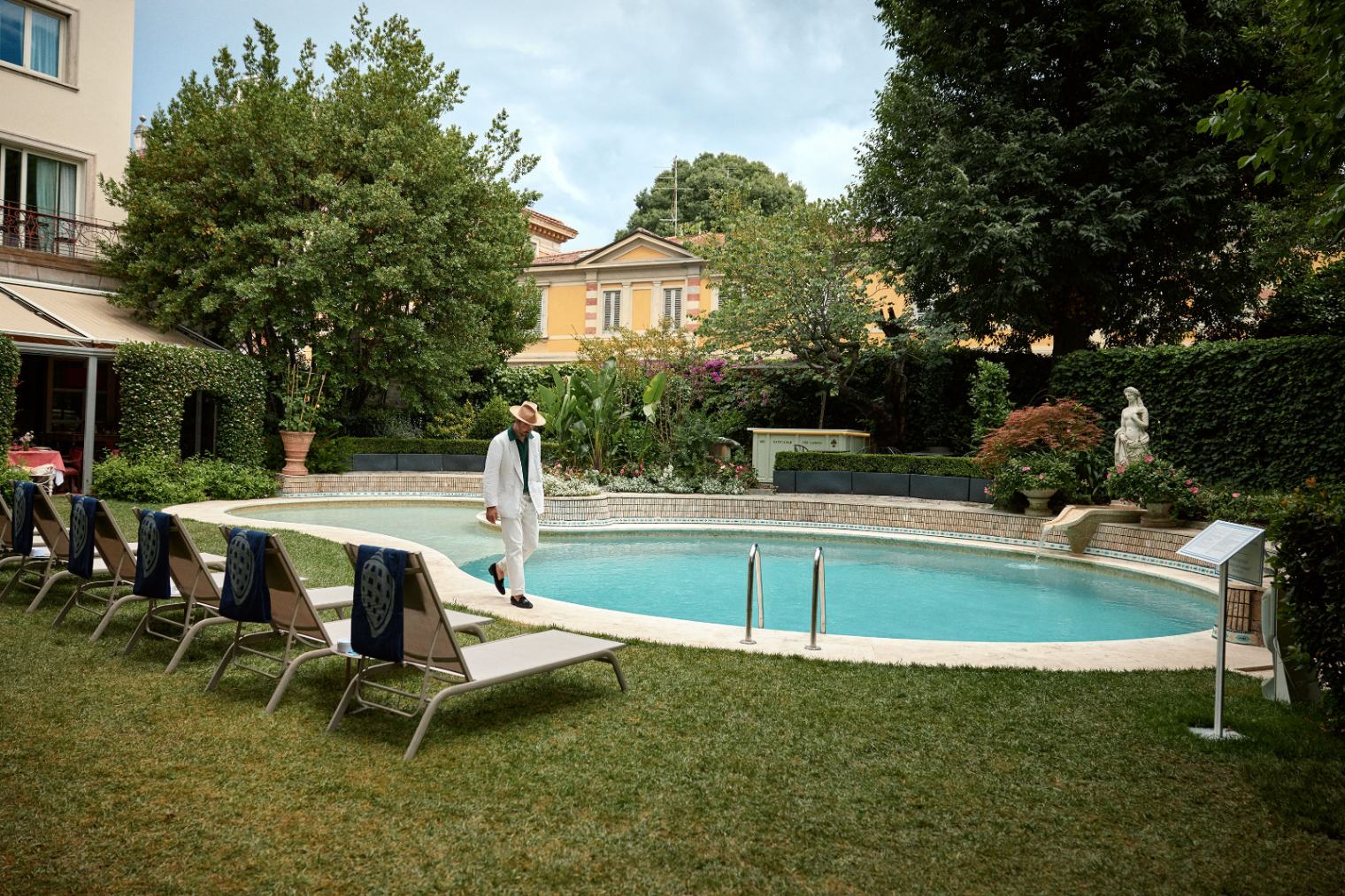 Stylish man in a white linen suit walking beside an elegant garden pool.