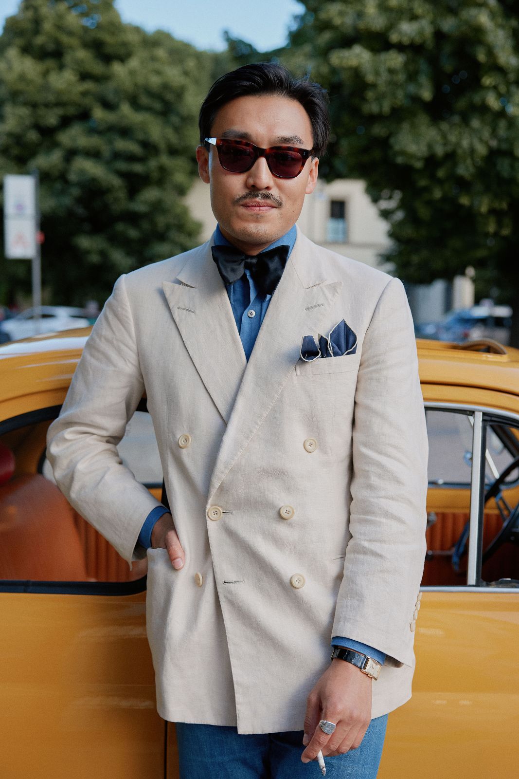 Well-dressed man in a beige linen suit by a vintage yellow car.