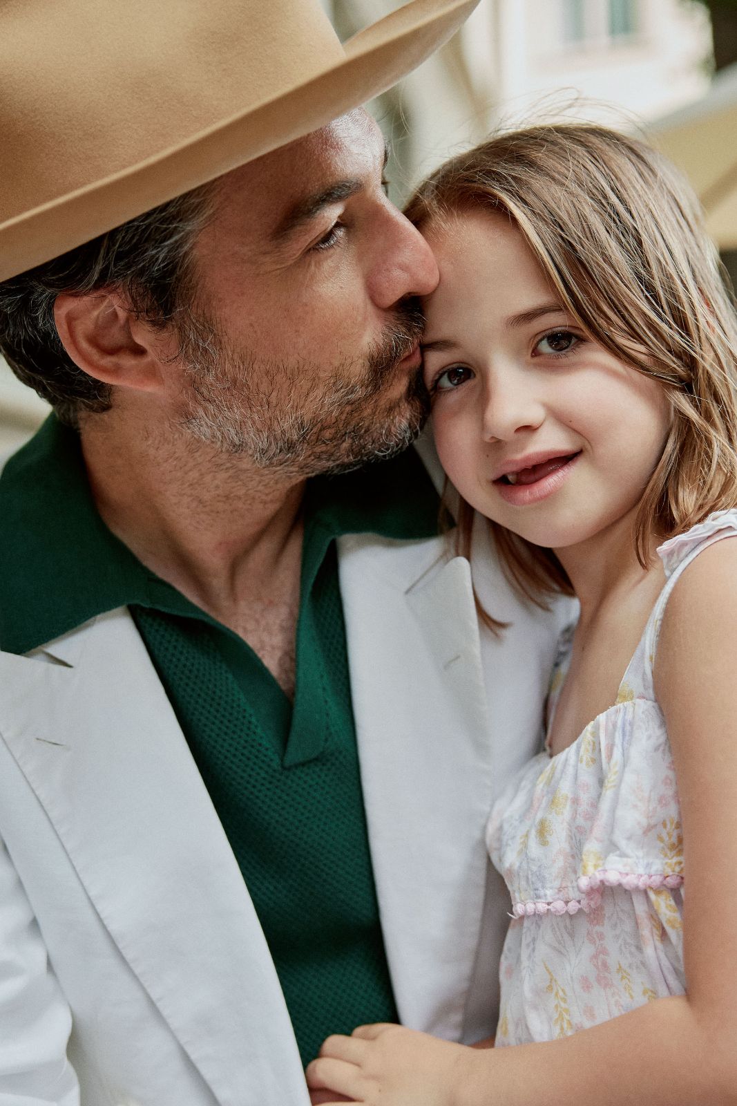 Tender moment as a father in a fedora kisses his daughter's forehead.