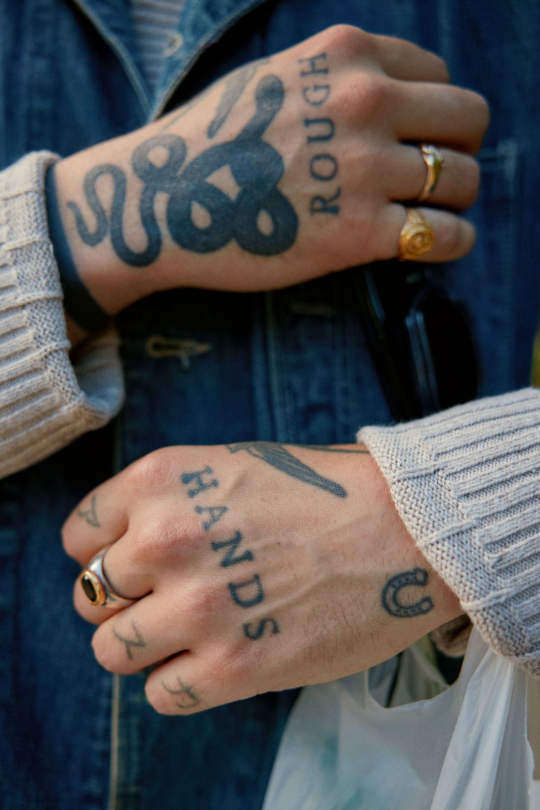 Crossed tattooed hands resting on a blue denim jacket in natural light.