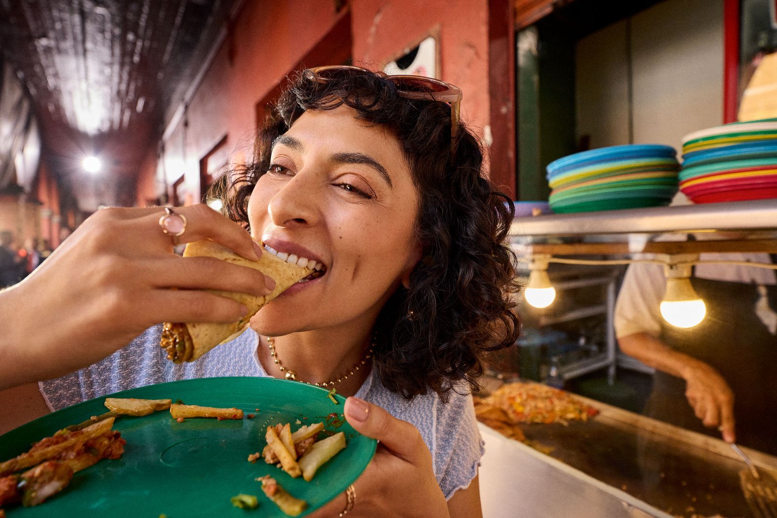 Smiling curly-haired woman enjoying fresh al pastor tacos at an energetic outdoor food stand.