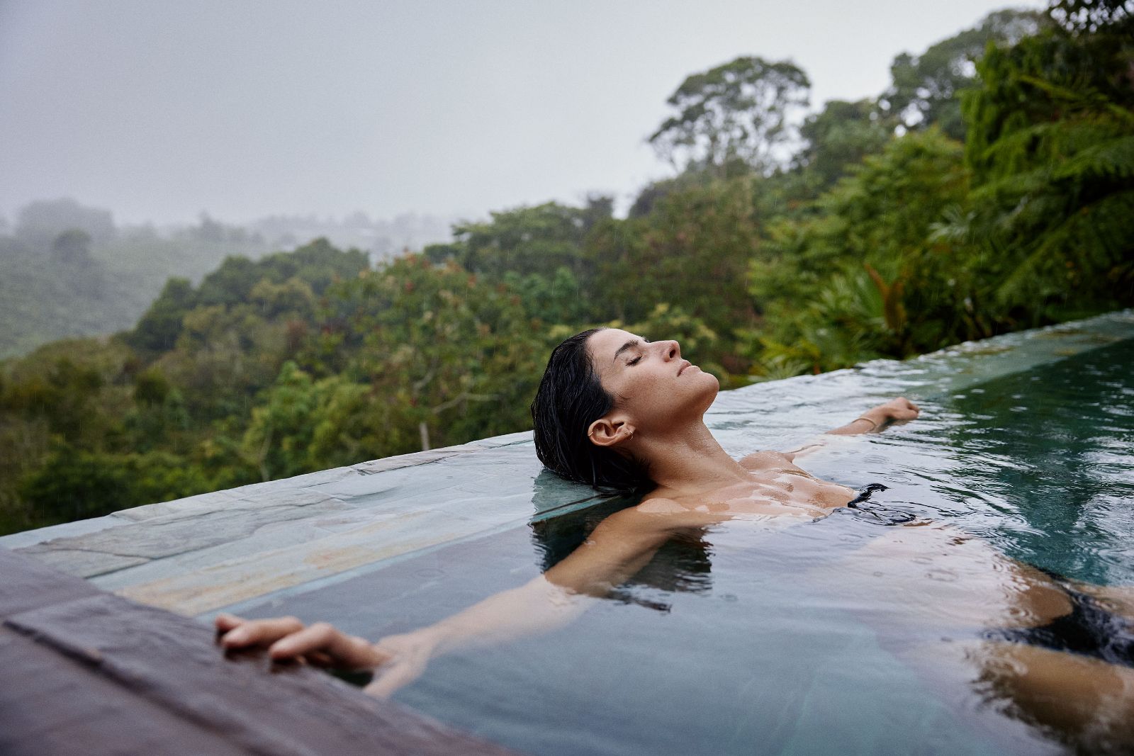 Kathy floating peacefully with eyes closed in an infinity pool overlooking a lush forest canopy.