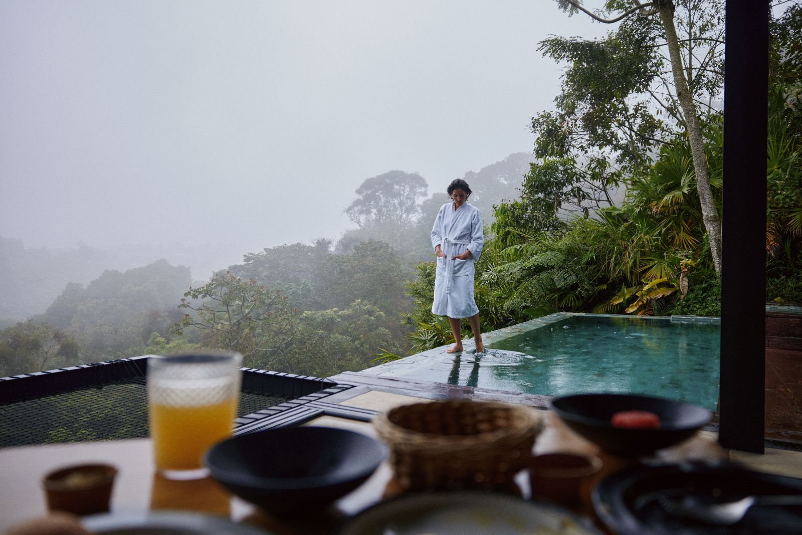 Kathy standing in a white robe at the edge of an infinity pool amidst misty mountains.