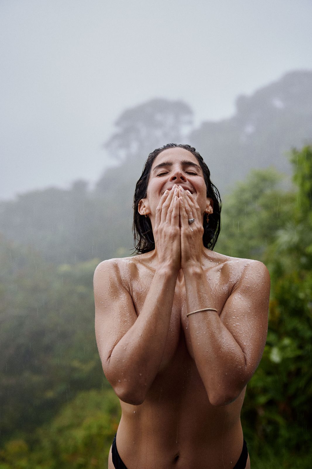 Close-up portrait of Kathy smiling with hands over her face while standing in tropical rain.