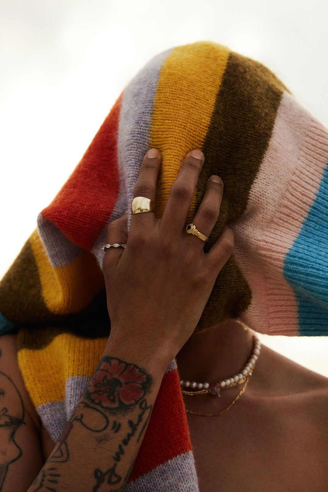 A hand with multiple rings rests gently against a colorful knit sweater worn loosely.