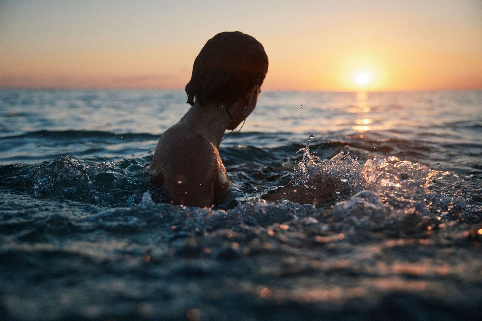 A woman splashes in the ocean while water droplets frozen mid-air catch golden sunlight.