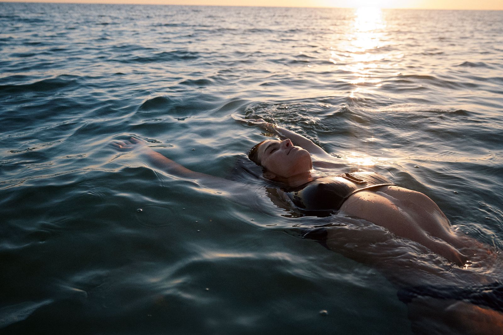 A woman floats calmly in the ocean as golden sunrise light illuminates the dark water.
