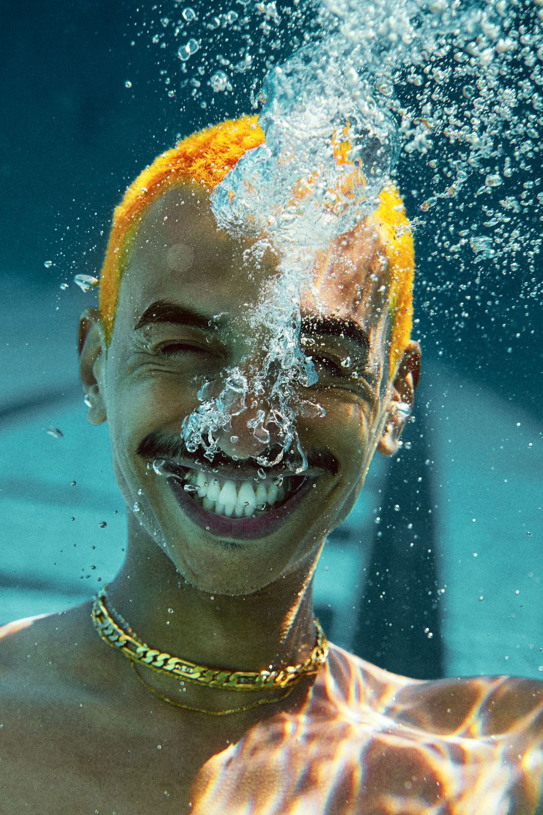Man with bright orange hair submerged underwater, smiling through a rush of silvery bubbles.