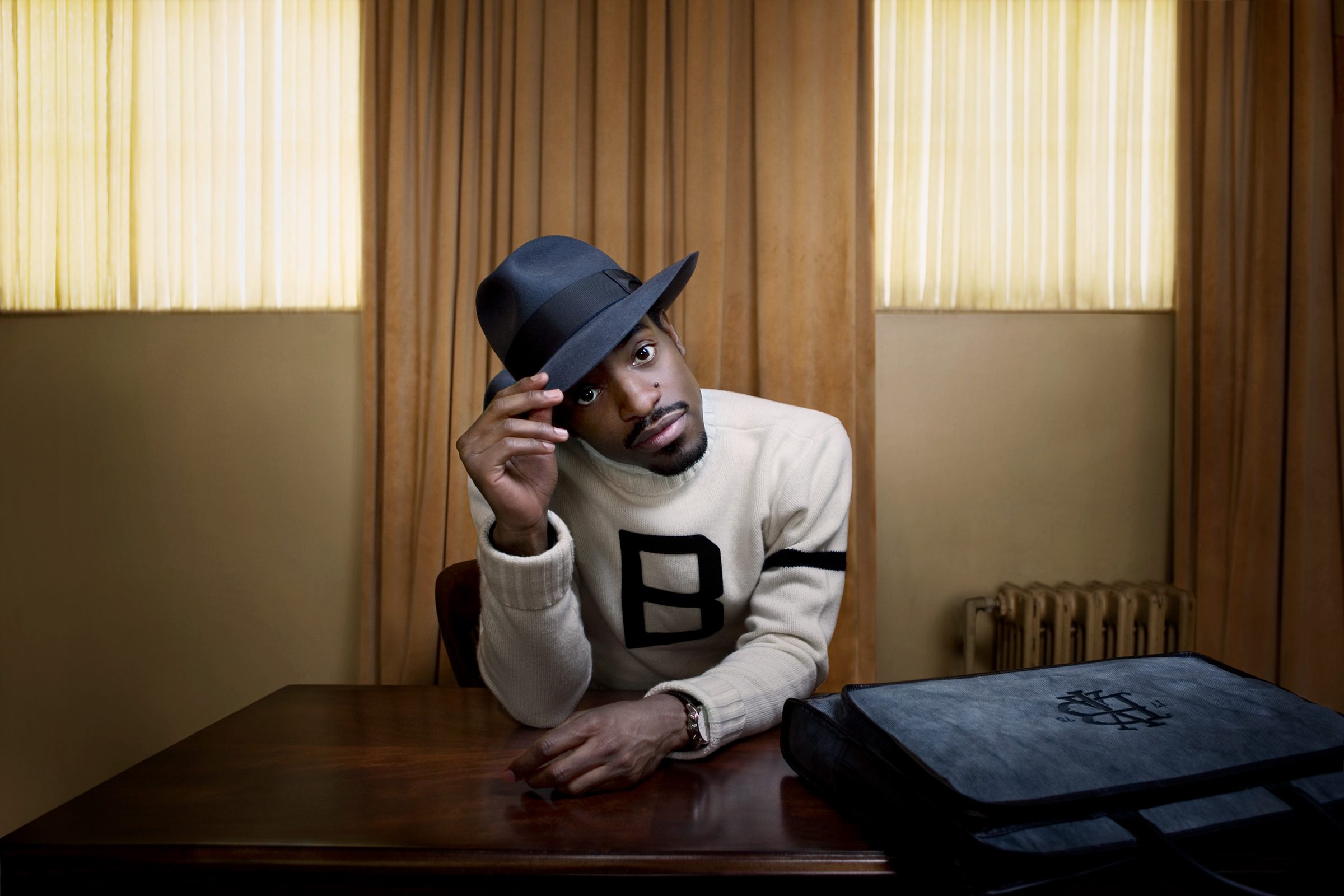 André 3000 tips his fedora at a wooden table, framed by amber curtains and a vintage radiator.