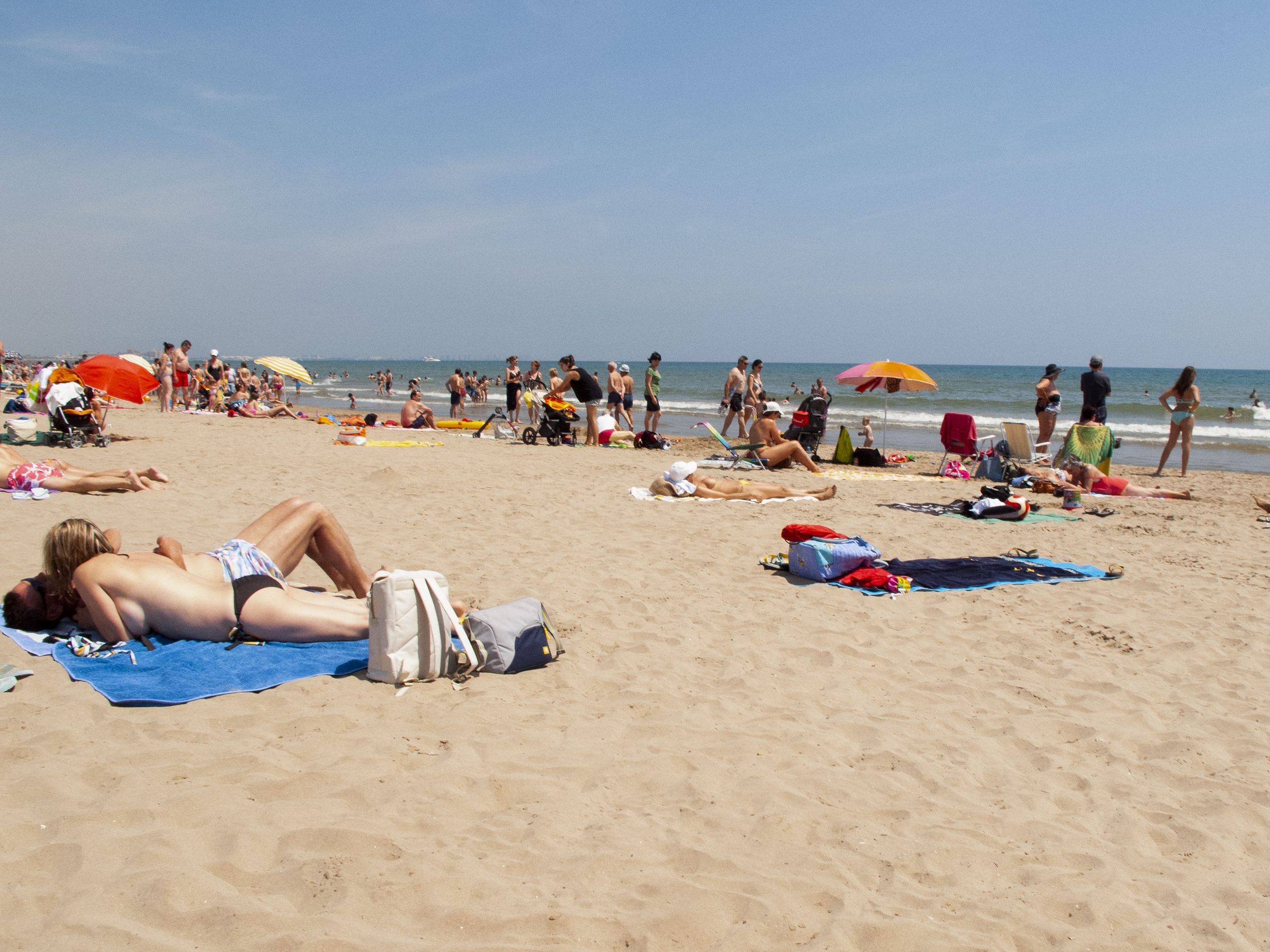 Sunbathers scatter across the packed sand, a man dozing on a blue towel in the foreground.