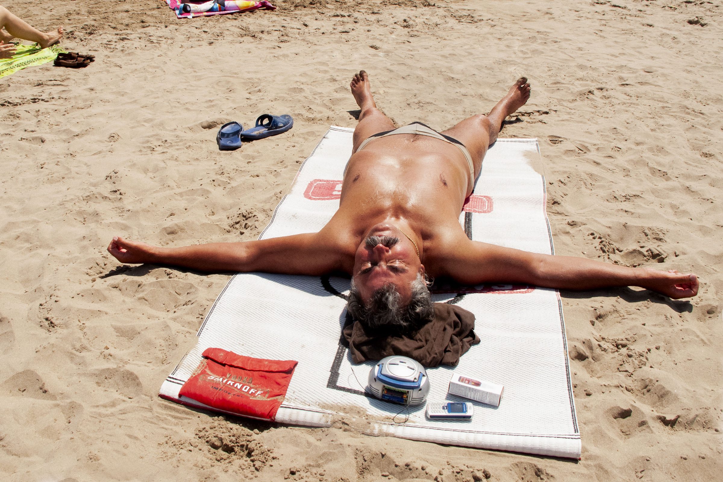 A sun-browned man lies spread-eagle on his towel, surrendered completely to the midday sun.