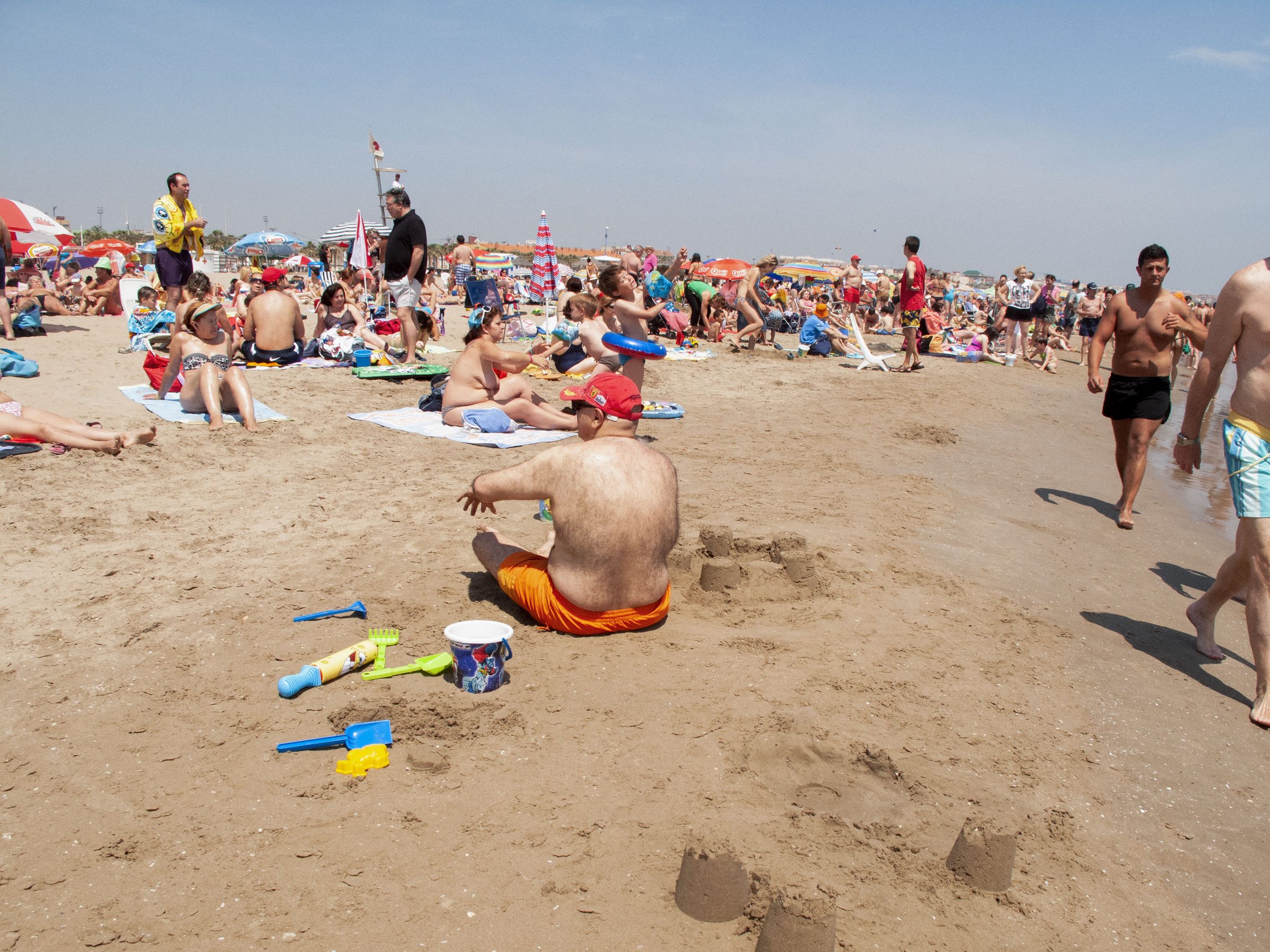 A sandcastle rises in the foreground as beachgoers stream past with buckets and spades.