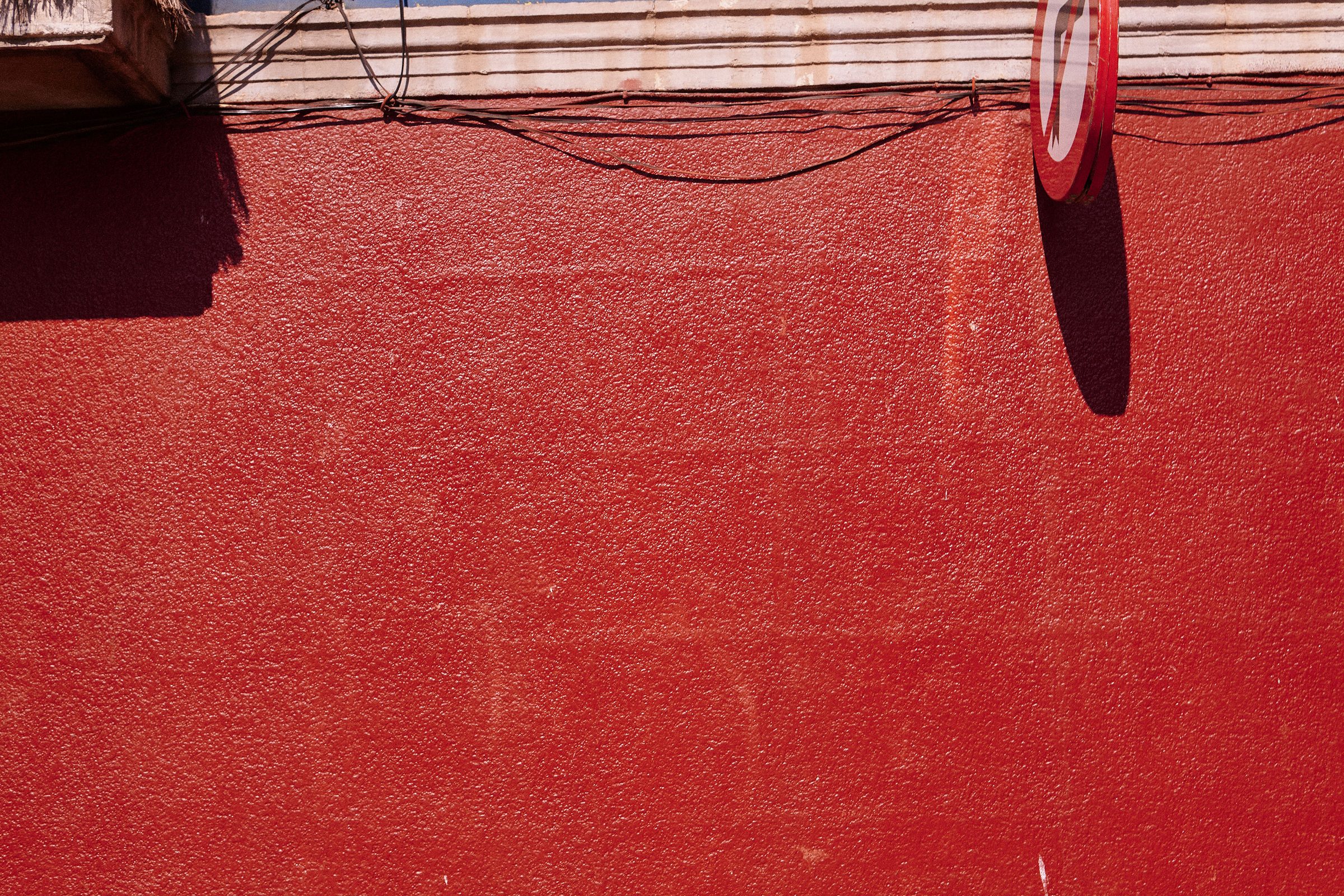Cable shadows draped across a sun-baked red wall beneath a traffic sign.