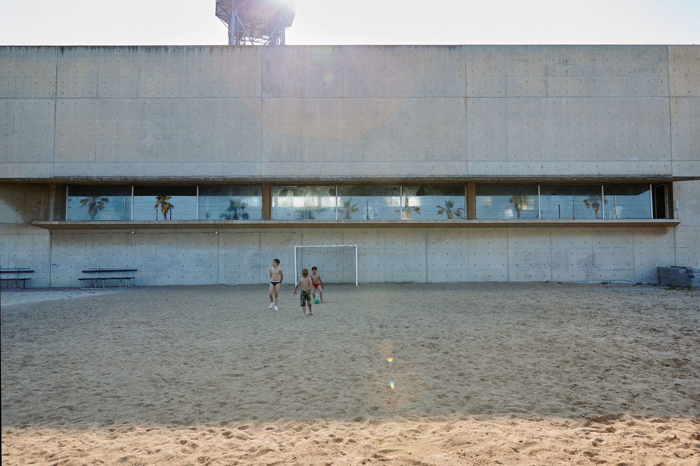 Two kids kick a ball around a goalmouth on a sand pitch beneath brutalist concrete stands.