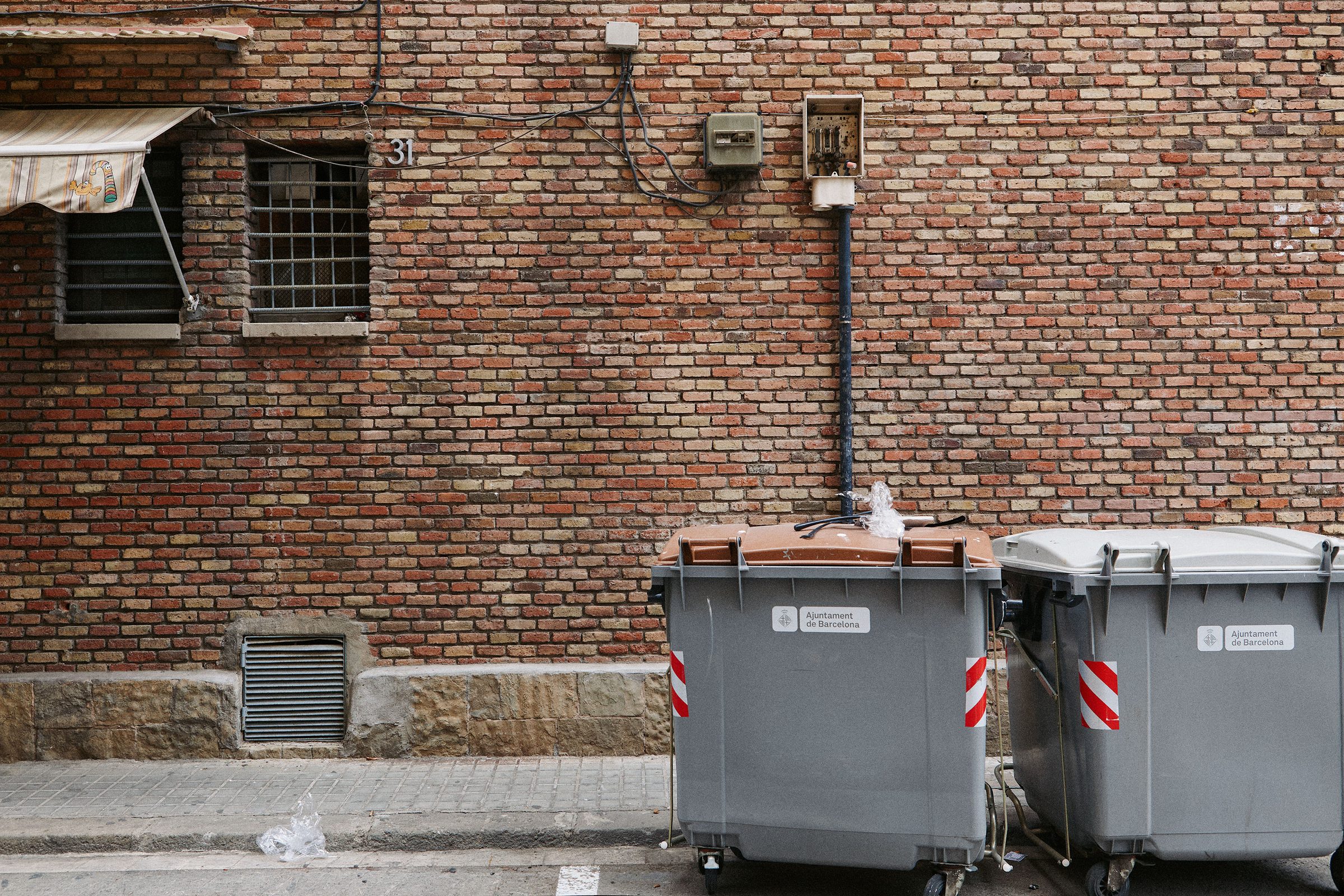 Grey dumpsters stationed against a patchwork brick wall in a back street.
